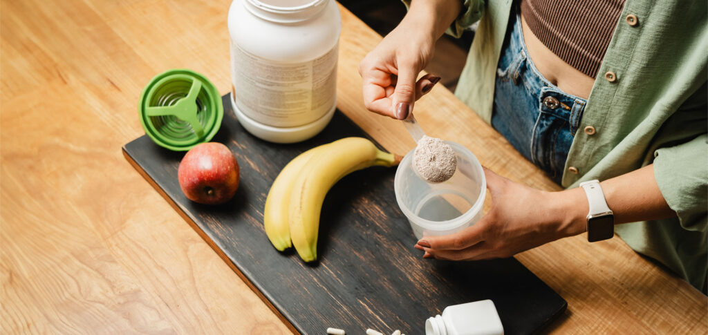 Woman in jeans and shirt with a measuring spoon in her hand puts portion of whey protein powder into a shaker on wooden table with white capsules, bananas and apple. Process of making protein drink.