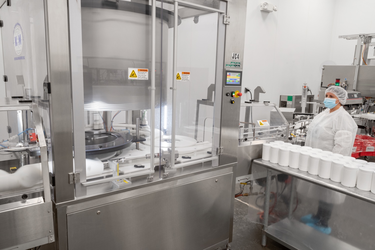 A worker in protective clothing operates a packaging jar capper machine